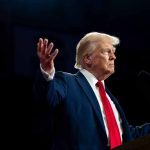 Person in suit gestures while speaking at podium.