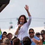 Person speaking with microphone, waving to outdoor crowd.