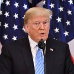 Man speaking at podium with American flags behind.