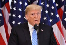 Man speaking at podium with American flags behind.