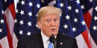 Man speaking at podium with American flags behind.