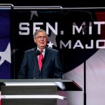 Man speaking at podium with American flag backdrop.