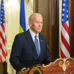 Man speaking at podium with flags in background.