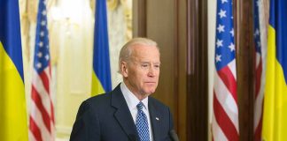 Man speaking at podium with flags in background.