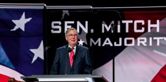 Man speaking at podium with American flag backdrop.