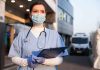 Nurse in scrubs and mask outside hospital holding clipboard
