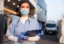 Nurse in scrubs and mask outside hospital holding clipboard