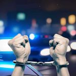 Handcuffed hands raised, police cars with lights on.