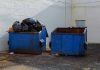 Two blue dumpsters, one overflowing with trash bags.