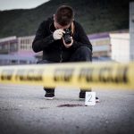 Brutal Murder Shocks Quiet Suburb Crime scene photographer behind police tape with evidence marker.