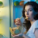 A woman eating a snack while looking at the camera, standing in front of an open refrigerator