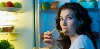 A woman eating a snack while looking at the camera, standing in front of an open refrigerator