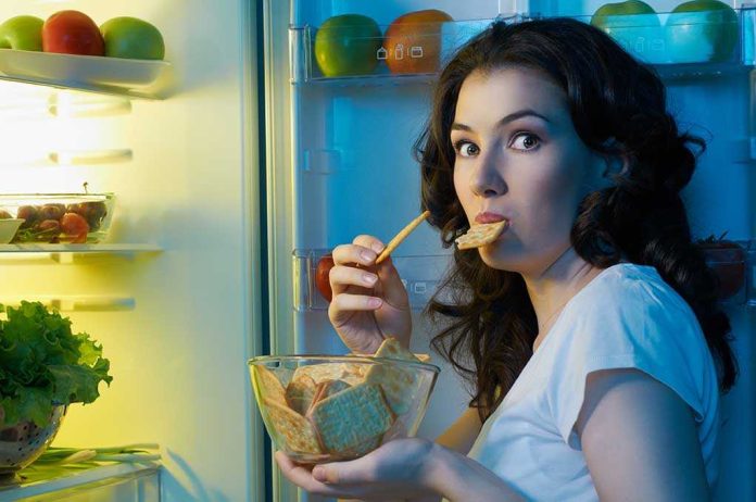 A woman eating a snack while looking at the camera, standing in front of an open refrigerator