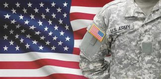 Soldier in camouflage uniform standing before American flag.
