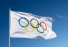 Olympian Murder Plot Shocks DOJ Olympic flag waving against clear blue sky.
