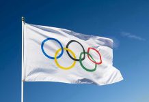 Olympian Murder Plot Shocks DOJ Olympic flag waving against clear blue sky.