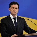 Zelensky’s Plane Targeted by Mystery Drones Man in suit speaking at podium with microphones.