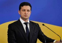 Zelensky’s Plane Targeted by Mystery Drones Man in suit speaking at podium with microphones.