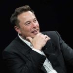 Musk’s God Bombshell Shocks Silicon Valley Man in suit smiling, resting chin on hand.