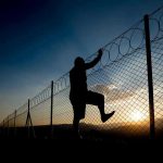 Silhouette of person climbing barbed wire fence at sunset.
