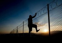 Silhouette of person climbing barbed wire fence at sunset.