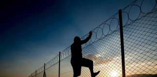 Silhouette of person climbing barbed wire fence at sunset.