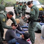 Law enforcement officers process a seated group outdoors