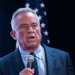 RFK Jr. Says Democrats Aren’t the Party They Once Were Man holding microphone, speaking with American flag behind.