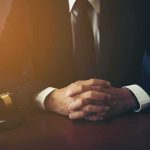 person in suit with hands clasped at a desk beside a gavel