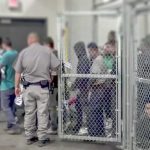 Hunger Strike? DHS Says No — Arrests Pile Up People standing inside chain-link holding area with an officer nearby