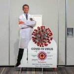 How Will History Remember Anthony Fauci? Cardboard cutout of doctor with COVID-19 virus info display