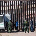 Spain’s Mass Illegal Crossings Receives Instant Pushback Border agents escort a group near a tall fence and a patrol van