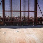 Ceuta Meltdown Exposes Legal Trap Crowd stands behind tall metal fence at border crossing
