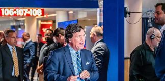 Tucker Carlson at a broadcast desk in a busy studio