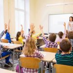 Students raise hands in a classroom while a teacher stands at the front