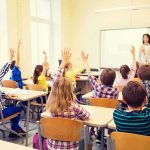 Students raise hands in a classroom with a teacher at the front