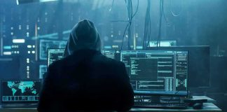 hooded person at multiple computer monitors in dim room