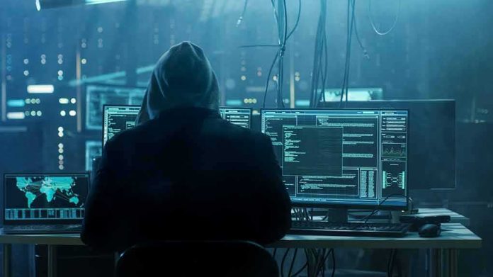 shutterstock_680075014 hooded person at multiple computer monitors in dim room