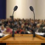 Cal State Professor DOXXES ICE Agent at Conference Podium with two microphones facing a blurred audience