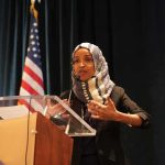 Democrats Cry ‘REVENGE’ Over Ilhan Omar Raid Ilhan Omar speaking at a podium with a microphone