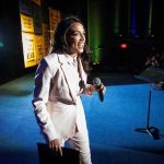 AOC Pours $1M Into Key Senate Race Woman in a light suit holding a microphone on stage