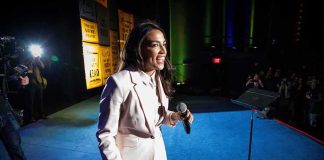 AOC Pours $1M Into Key Senate Race Woman in a light suit holding a microphone on stage