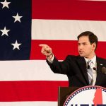 Rubio Tells the West to Stop APOLOGIZING Speaker in suit pointing during speech before large American flag